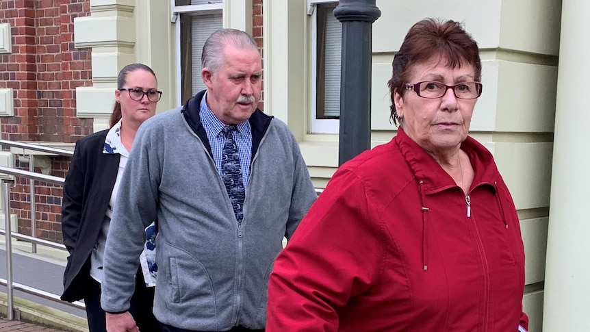 A woman in a red sweater looks at the camera as she leaves a court building followed by a man and another woman
