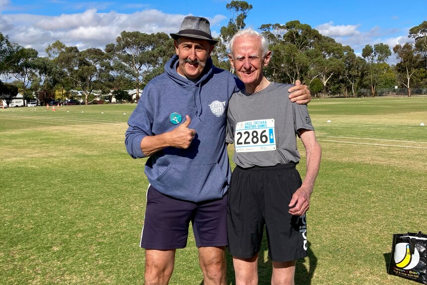 A man giving a thumbs-up while an elderly man wears an athletics bib with a number on it