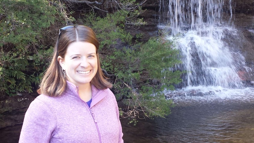 Erin Davidson smiles in her pink jacket while standing next to a brown creek in front of a waterfall.