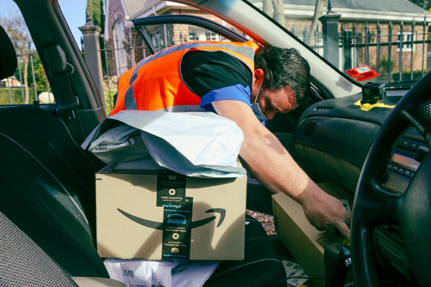 A man in an orange high-vis vest bends over to reach a package in the foot well on the passenger side of his car.