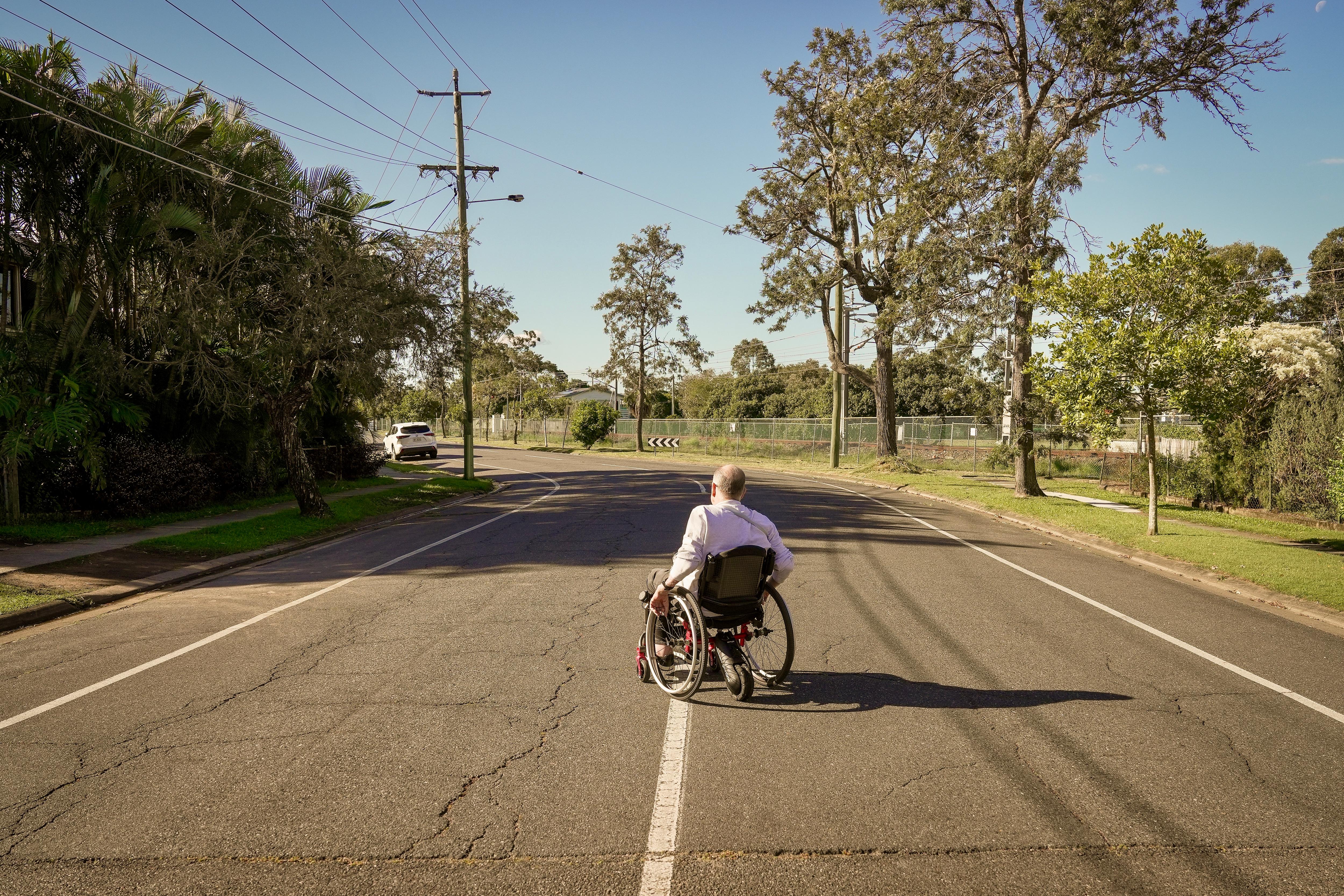 A middle aged white man with short grey hair. He's in a wheelchair, stationary in the middle of a surbuban road