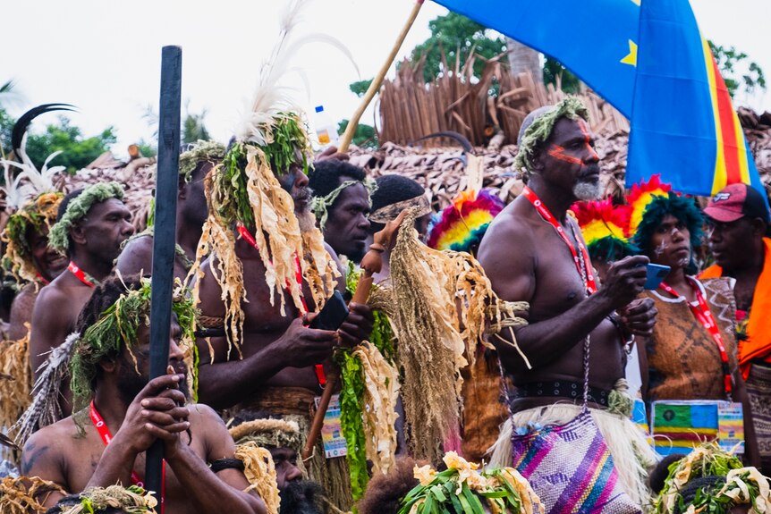A group of Melanesian cultural performers wearing traditional woven wear and holding a blue flag