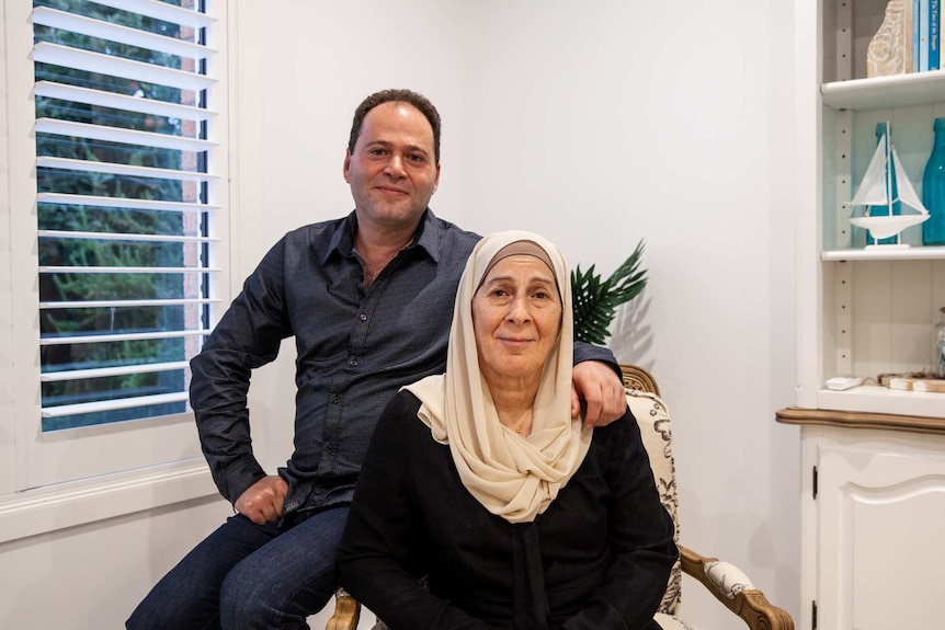 You view a middle aged man leaning against a chair, with an elderly woman in a Hijab sitting next to him.