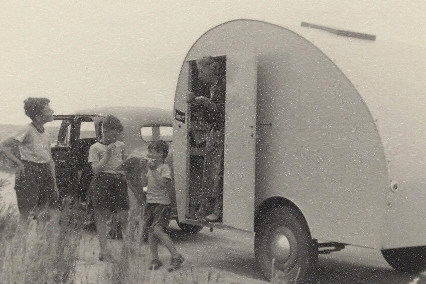 Black-and-white photograph of a woman leaning out the door of a caravan, with three sons standing nearby.