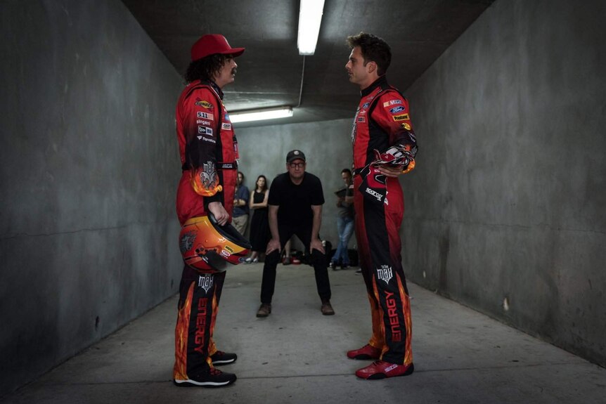 Still image of Seth MacFarlane and Sebastian Stan dressed in NASCAR attire standing in a narrow concrete corridor.