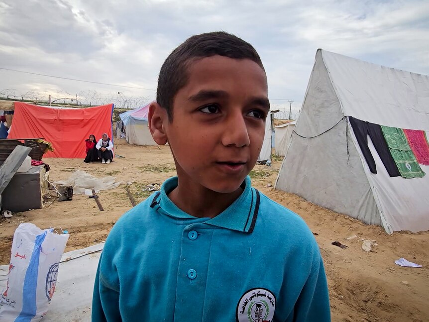A young boy wearing blue collared shirt stands in front of tents