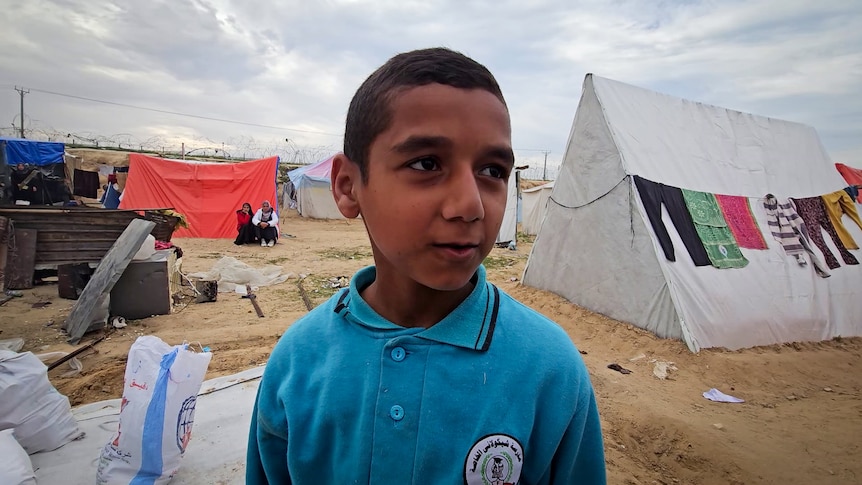 A young boy wearing blue collared shirt stands in front of tents