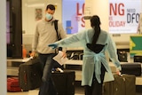 A man wearing a face mask and carrying luggage is directed by a woman in PPE near a baggage carousel at Perth Airport.