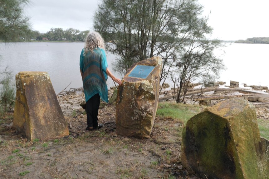 Woman with long grey hair stands between three large stones looking out across the water, hand on the stone with a plaque.