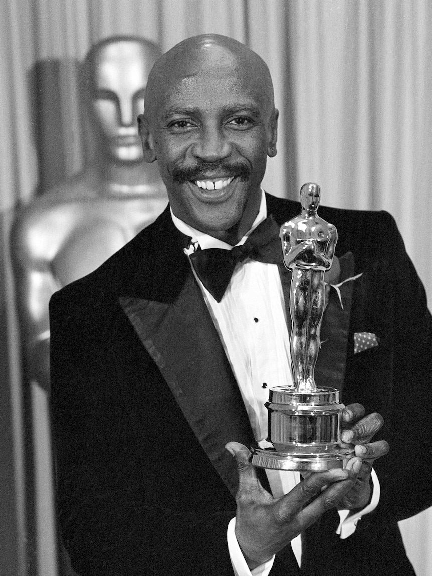  Louis Gossett Jr. smiles as he holds up an Oscar in a black and white photo.