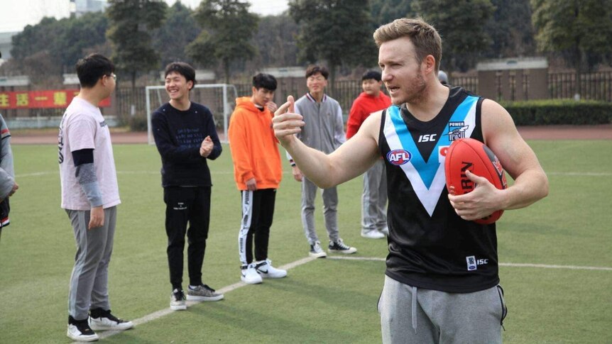 Australian expat Tyson Murphy holds an AFL football in front of several Chinese students at a football field.