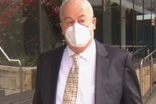 An older man in a suit, wearing a mask, walking outside a court complex.