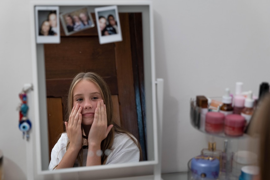 A mirror showing the reflection of a young Caucasian girl with long blond hair touching her face sitting near skincare products.