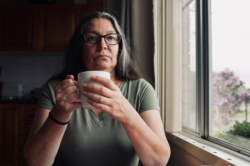 A woman with dark hair and glasses holds a cup of tea and looks straight into the camera.