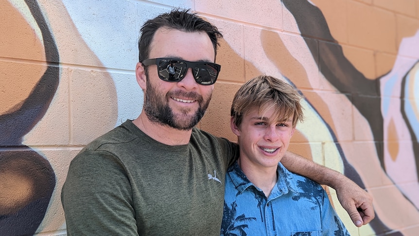 A man with sunglasses and a green t-shirt has his hand around his son in a tropical shirt in front of a mural in Alice Springs