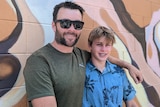 A man with sunglasses and a green t-shirt has his hand around his son in a tropical shirt in front of a mural in Alice Springs