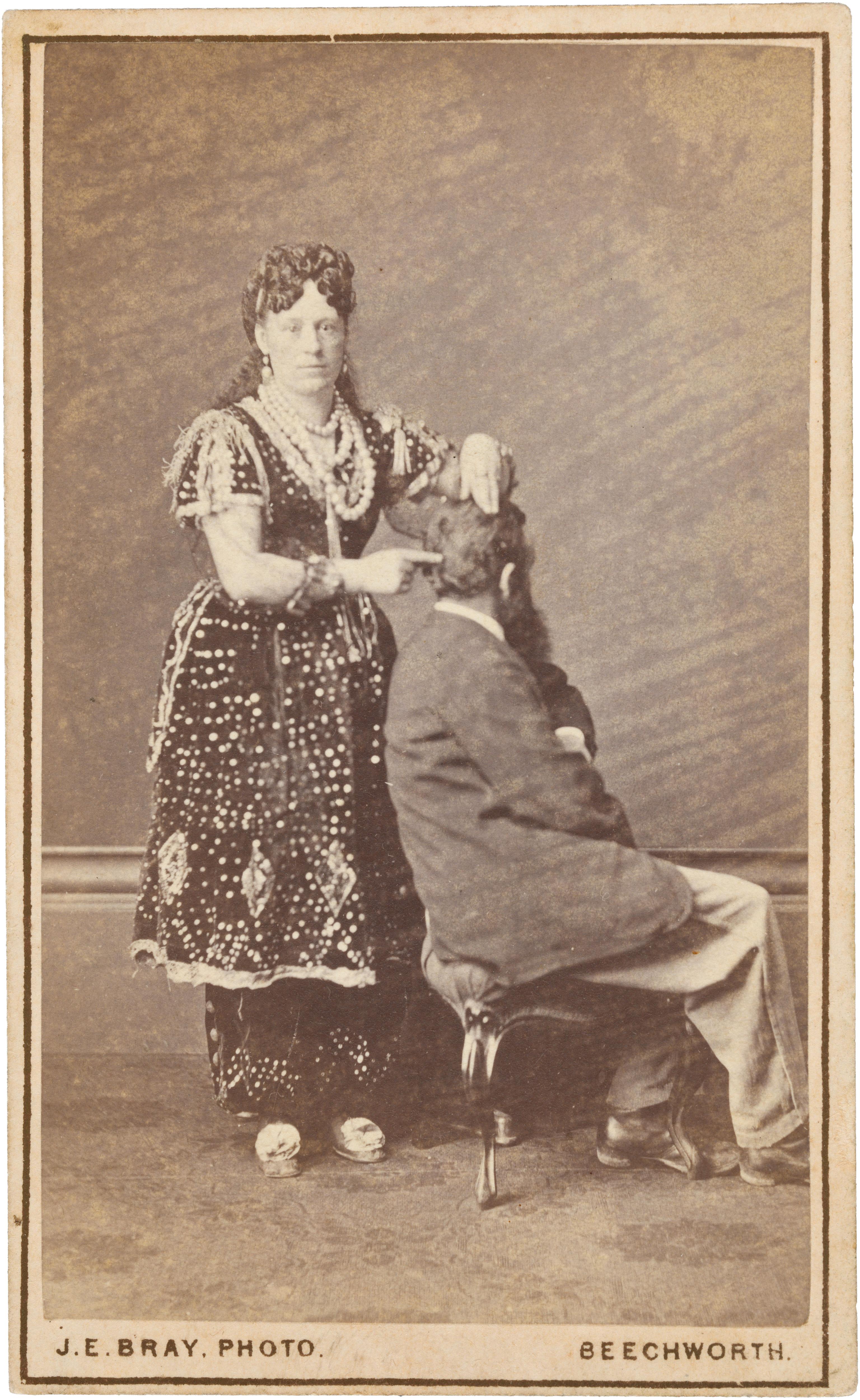 A late 1800s sepia photograph of a woman in a bejewelled dress pointing at a sitting man's head