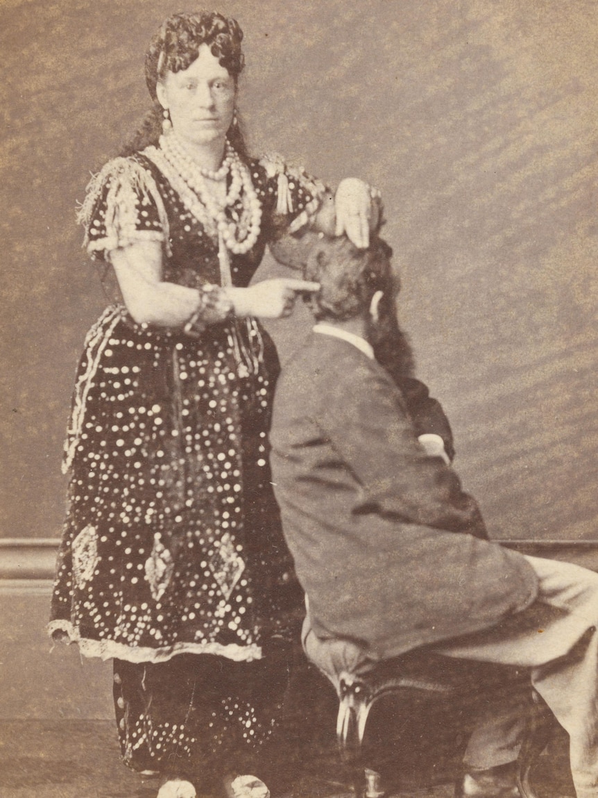 A late 1800s sepia photograph of a woman in a bejewelled dress pointing at a sitting man's head