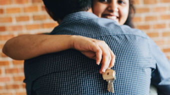 A smiling woman hugs a man while holding a keys on a keyring with a wooden house.