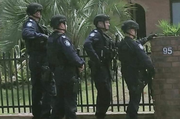 A group of four male police officers in combat gear stand at front of entrance of brick house.