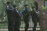 A group of four male police officers in combat gear stand at front of entrance of brick house.