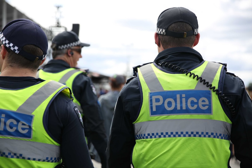 Three male police officers wearing high vis yellow vests with their backs to the camera.