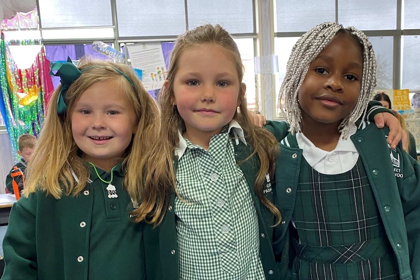Three primary school girls in green uniforms stand with their arms around each other's shoulders inside a classroom.