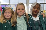 Three primary school girls in green uniforms stand with their arms around each other's shoulders inside a classroom.