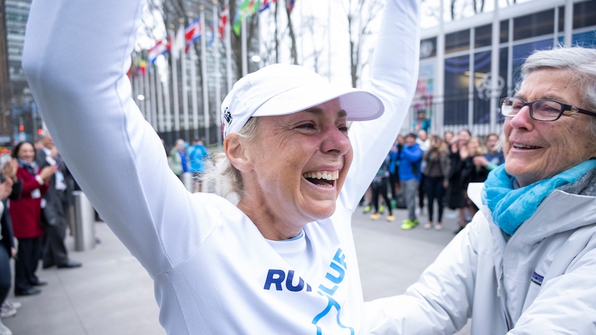 A woman wearing white top and white cap smiles and raises her hands in joy. 