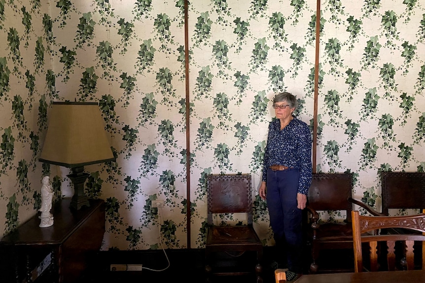 A woman stands in  a room with wallpaper featuring green vines.
