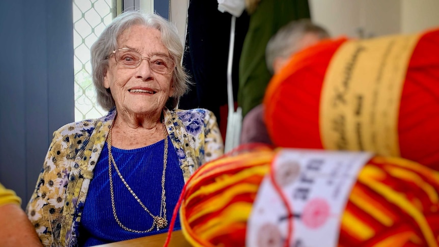 An elderly woman sits at a table filled with balls of wool.