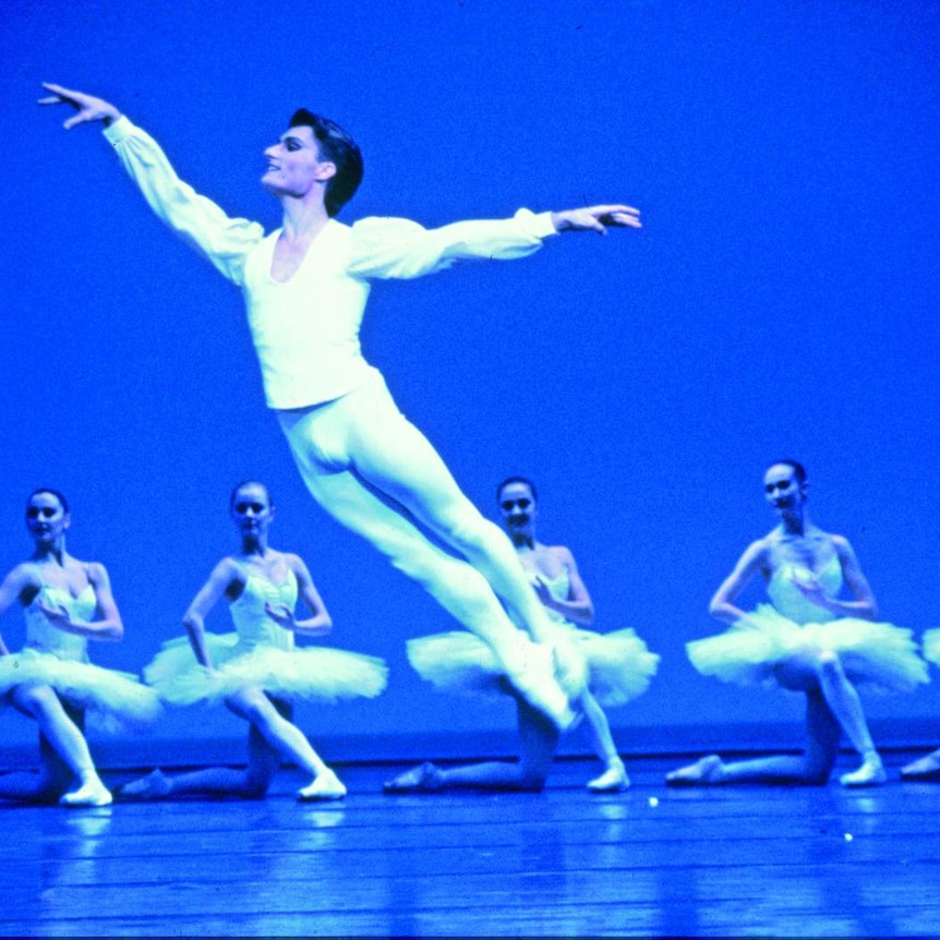 David McAllister in white leggings and blouse, jumping through the air with arms outstretched. Six ballerinas behind him