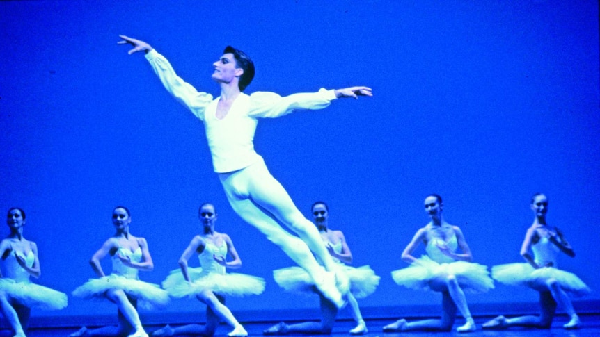 David McAllister in white leggings and blouse, jumping through the air with arms outstretched. Six ballerinas behind him
