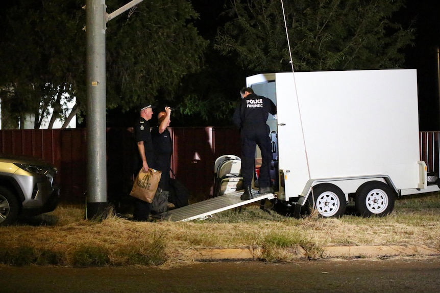 Police forensic officers stand at the rear of a white box trailer on a street verge at night.