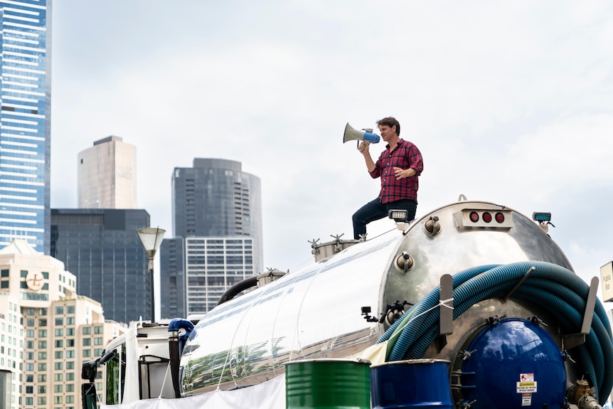 Man holding a megaphone standing on top of an oil tanker in a city location.