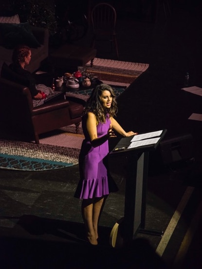 A woman in a purple dress speaks on stage behind a lectern. 