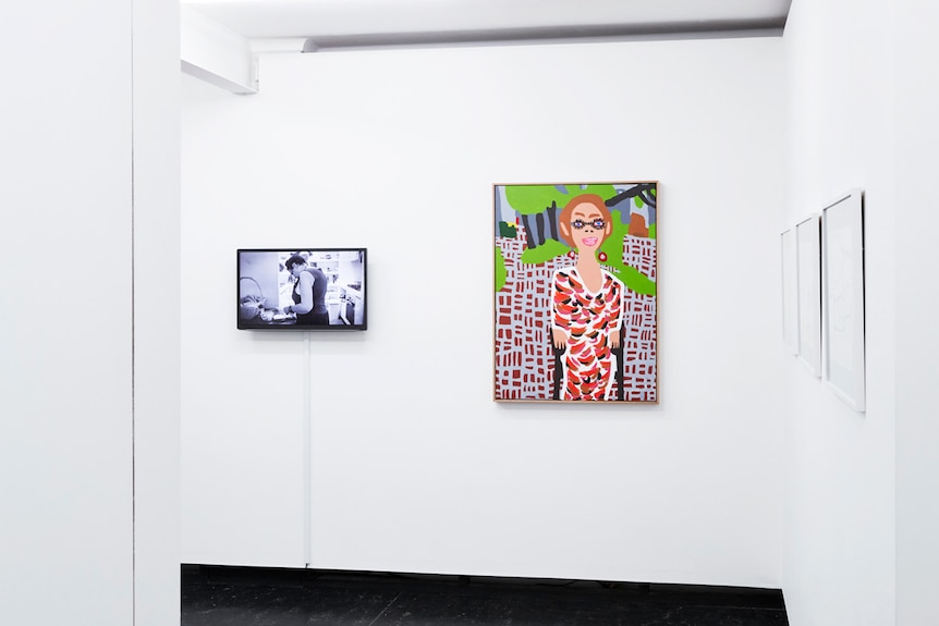 A black and white film on a video screen and a brightly coloured painted portrait hang side by side in a white gallery space.