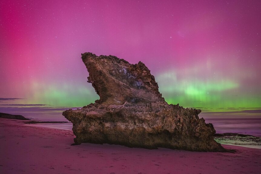 A pink and green aurora behind a rock. 