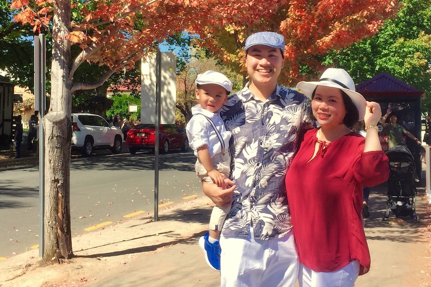 A family of three stand under an autumn tree wearing hats.