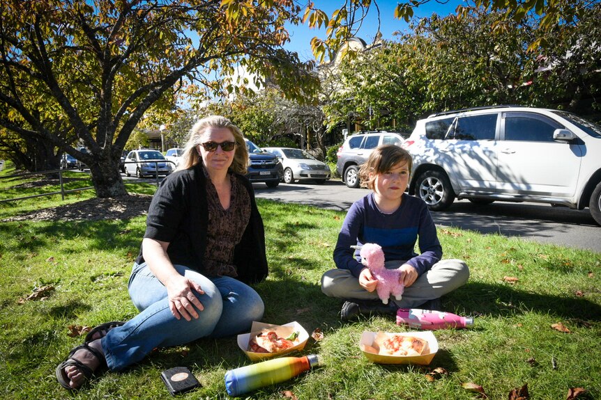 A woman and a child sit on green grass with cars passing in the background.