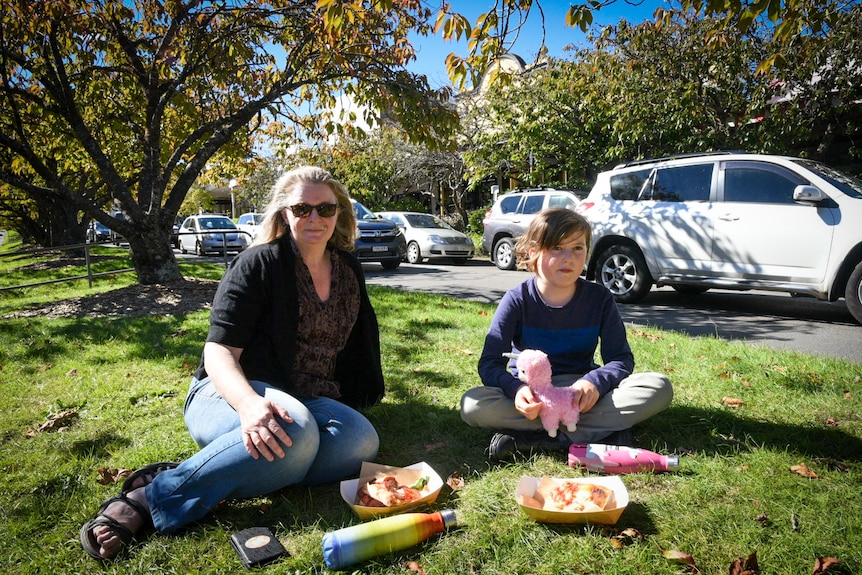 A woman and a child sit on green grass with cars passing in the background.