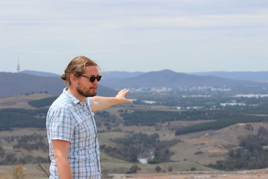 Man with hair pulled back in dark glasses and checked short-sleeved shirt pointing across distant landscape