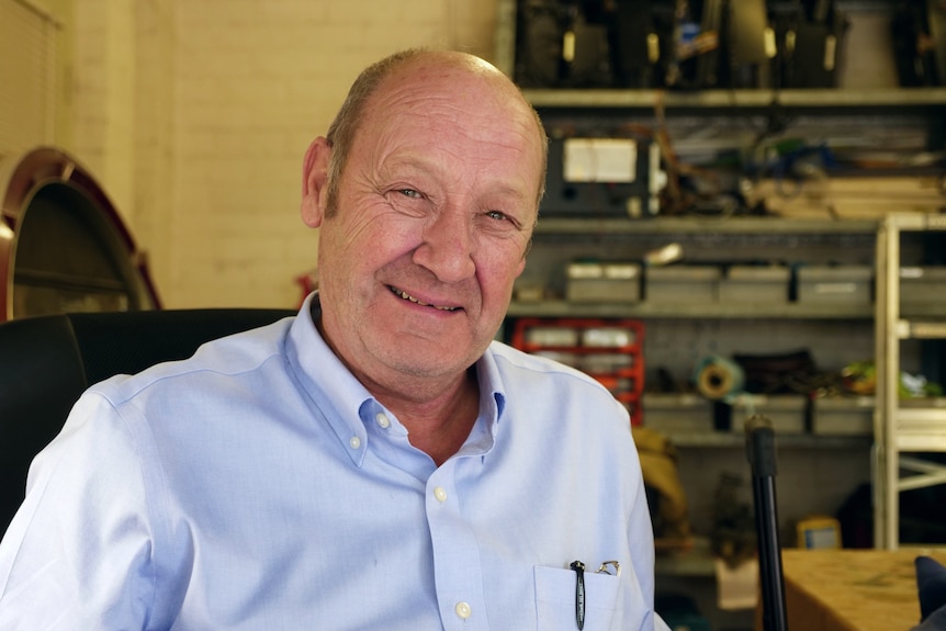 An older man with a receeding hair line and a blue shirt stares at the camera smiling