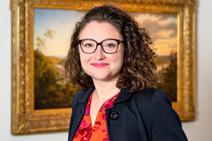 A 35 yr old professional woman stands smiling in front of an old landscape painting in a gallery