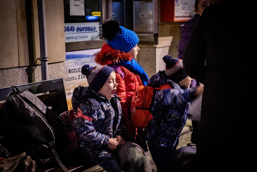 Three children sit on a bench, one yawns. All are in winter coats and beanies