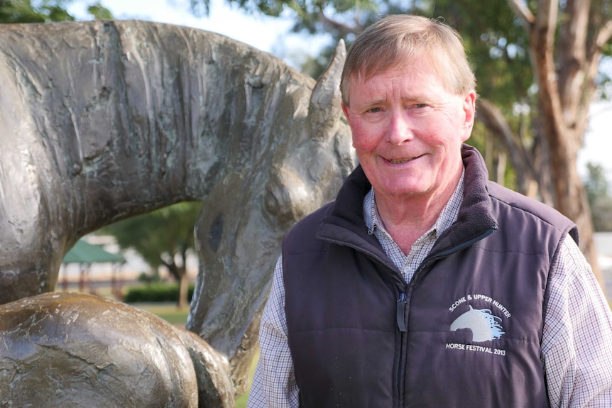 Elderly gentleman in checked shirt and jacket standing in front of a horse statue outside in a park.