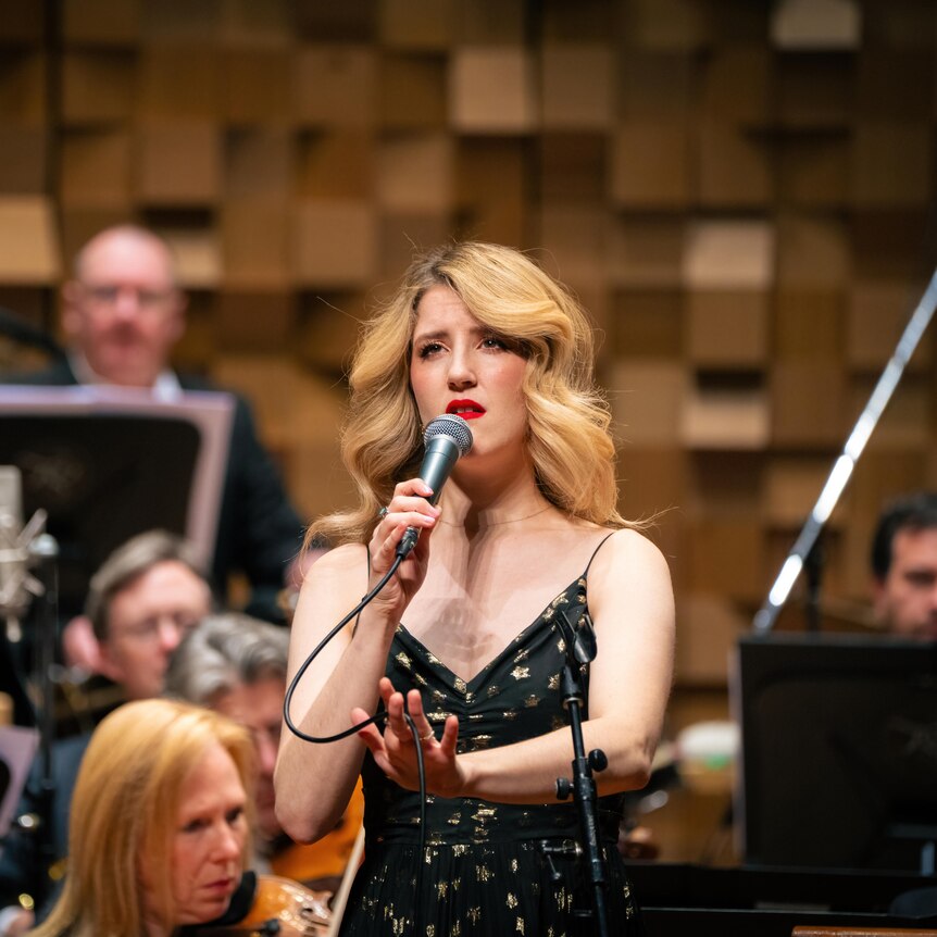  Singer Olivia Chindamo wears a black dress, she is holding a microphone. The orchestra is behind her.