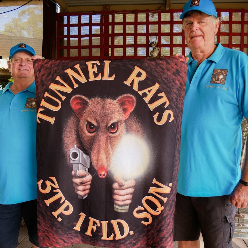 Two men standing, holding up a towel with tunnel rats and 3TP. 1FLD. SQN. written on it with a picture of a rat holding a gun.