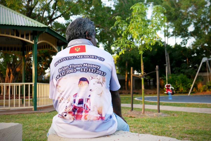A man sits on a concrete block and looks out at a park. He wears a shirt that reads 'in loving memory of Brian Flynn Massey'