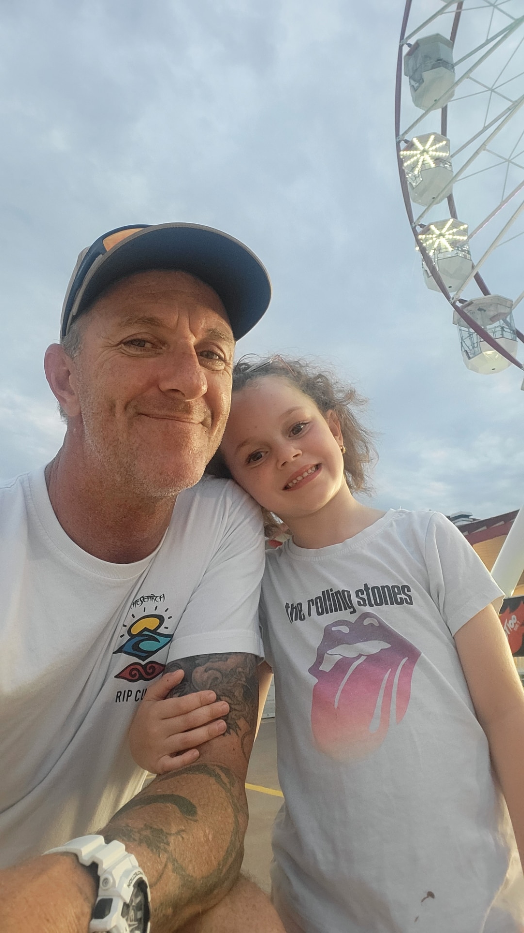 A man, white, with a little girl smiling softly at a camera with a ferris wheel in the background.